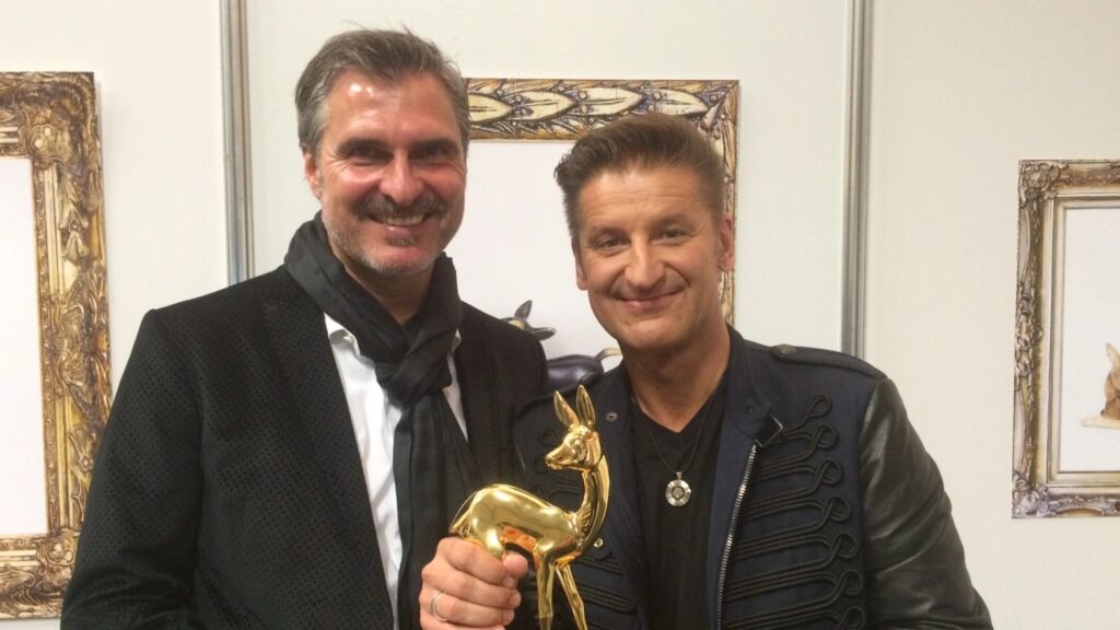Zwei Männer lächeln in einem Innenraum; einer hält eine goldene Hirschfigur, im Hintergrund sind gerahmte Bilder zu sehen.