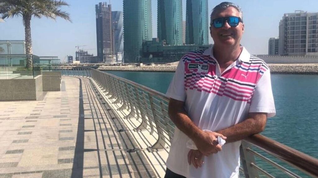 Mann mit Sonnenbrille lehnt an Geländer an einer Promenade am Wasser, im Hintergrund Hochhäuser und Palmen.