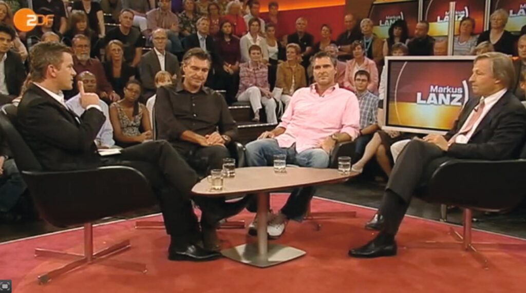 Studioaufnahme einer Talkshow: vier Männer sitzen in Sesseln um einen niedrigen Tisch, Zuschauertribüne im Hintergrund, Bildschirm mit Sendungslogo sichtbar.