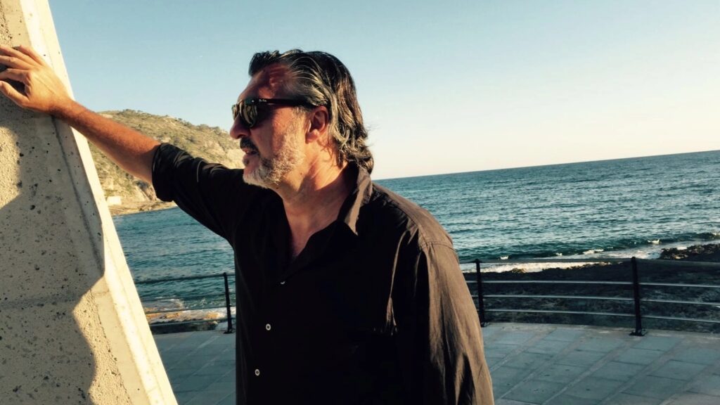Erwachsener Mann mit Bart und Sonnenbrille in dunklem Hemd lehnt an einer Mauer am Meer.
