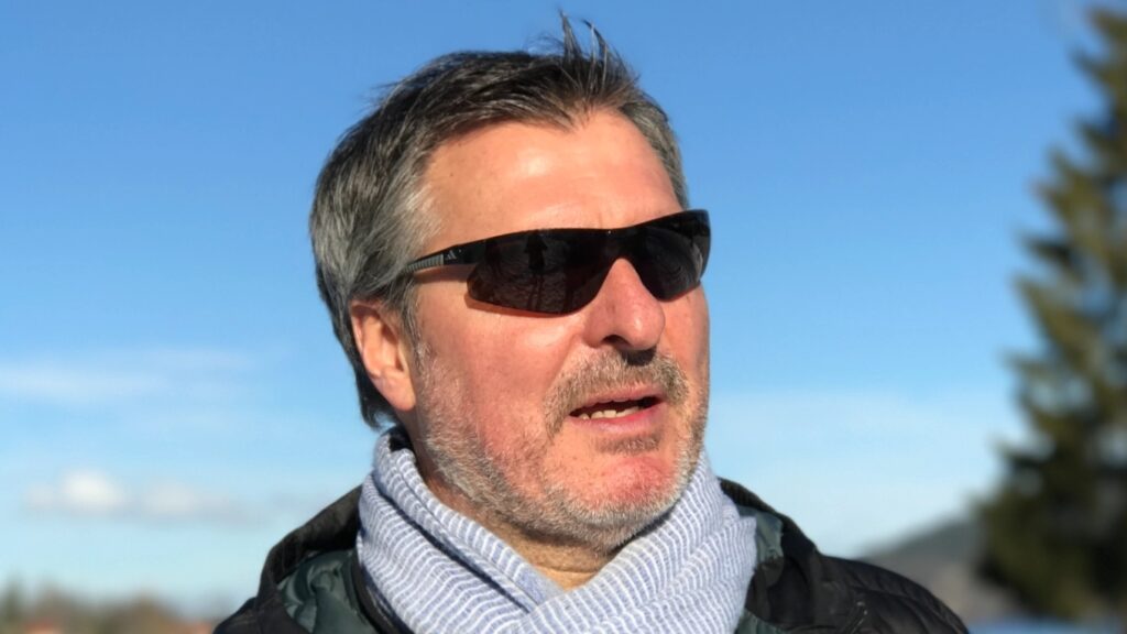 Porträt eines erwachsenen Mannes mit grauen Haaren, Sonnenbrille, Schal und Jacke vor blauem Himmel.